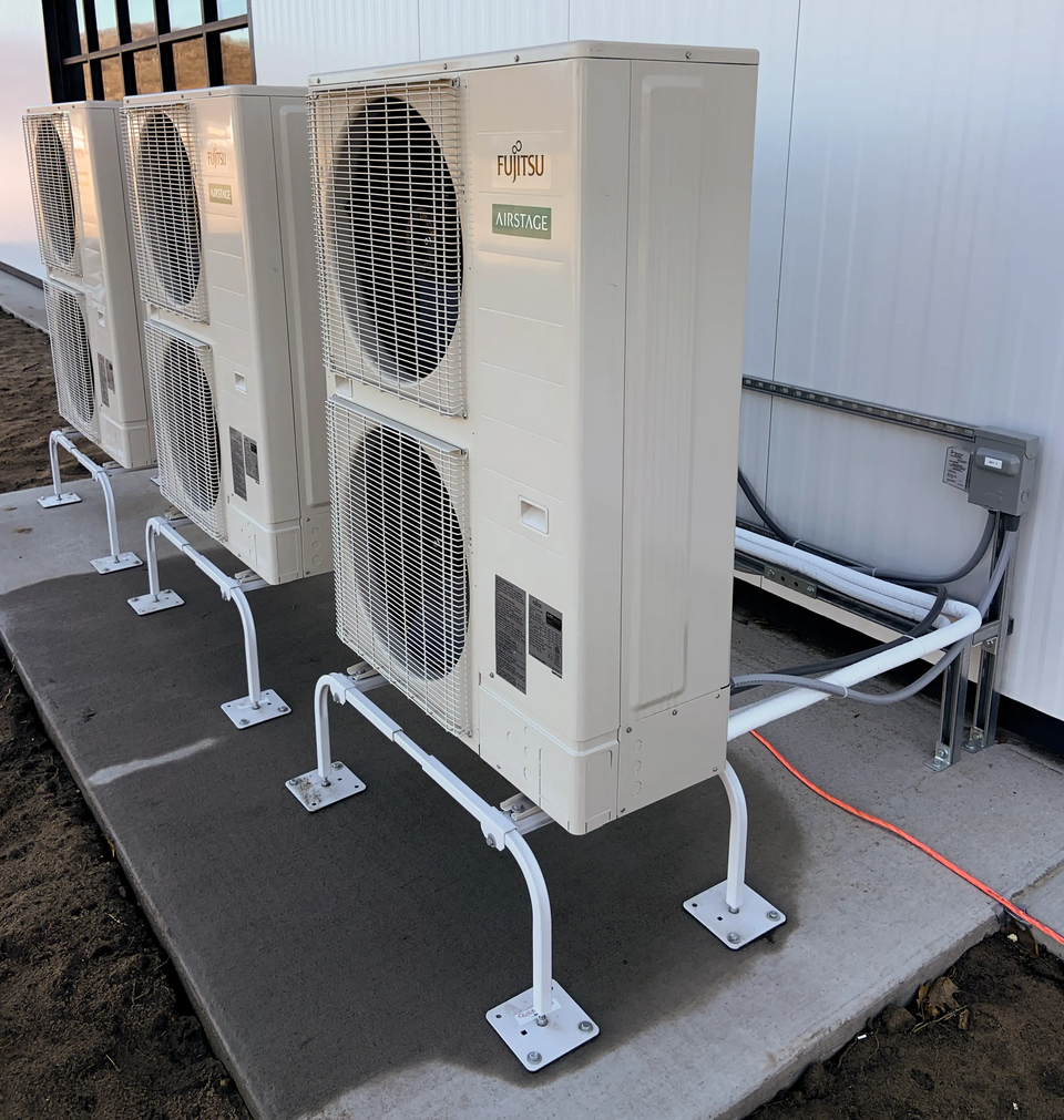 A diagonal shot of three heat pumps, each with two vertically stacked fans, mounted on a tubular steel base on a flat surface. White tubing emerges from the back of the closest unit. Behind the pumps, galvanized brackets secure cables and pipes to a tall white wall, and a multi-paned black window glows with yellow light from inside the building.