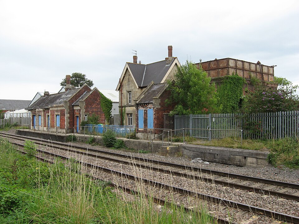 Former Charfield railway station building with boarded windows, disused platform, and metal water tank alongside active railway tracks in South Gloucestershire.
