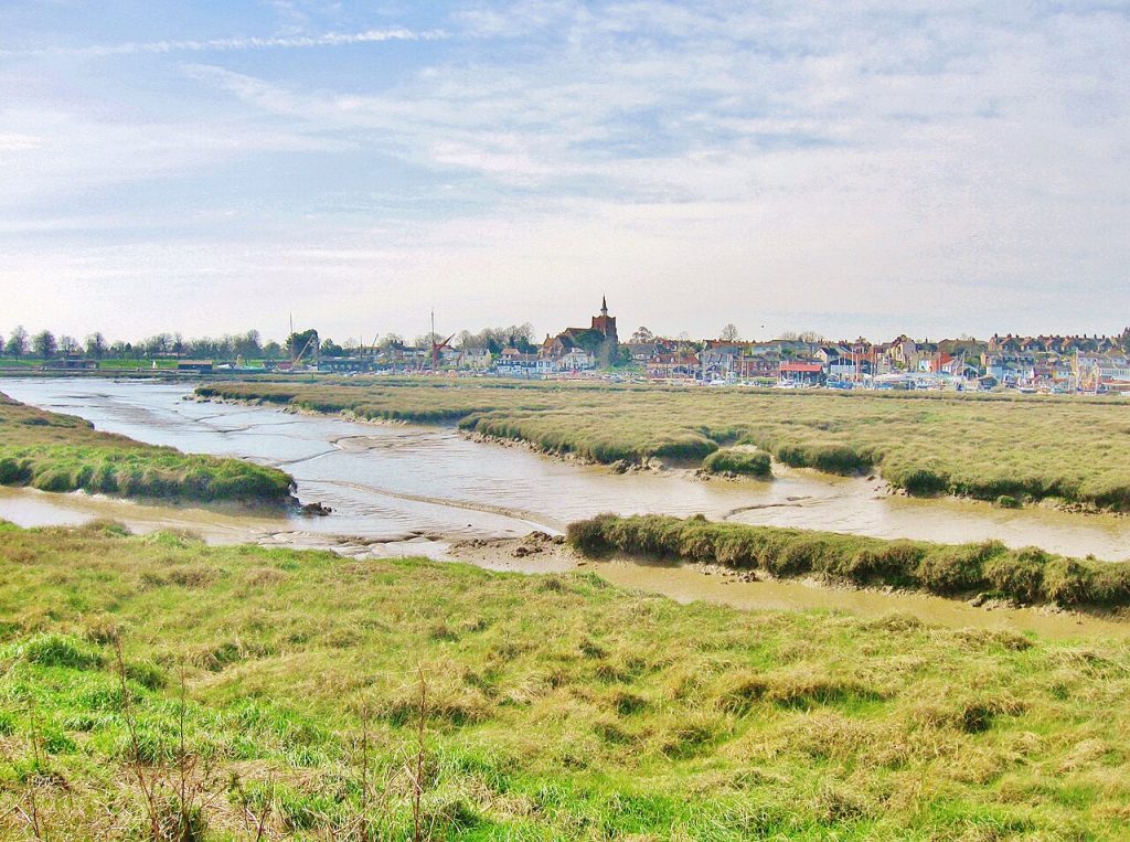 River Roach marshland at low tide in Essex, featuring exposed mud banks with visible drainage patterns, a marina with boat masts, and a distant town with a square church tower.