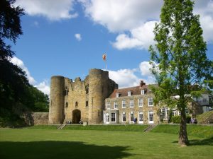 Tonbridge Castle in Kent featuring a medieval stone gatehouse on the left attached to a two-story Georgian mansion on the right, set behind a wide lawn under a clear blue sky.